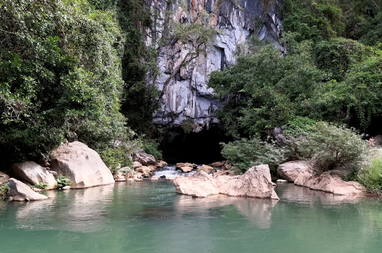 The river entrance of Kong Lor cave: a dark opening at the foot of a forested limestone cliff above a pool of clear green water